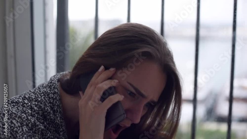 Close-up of depressed woman is talking over the mobile phone, yelling and crying sitting on the floor by the window with rain falling outside. Lonely woman in despair with the suicide thoughts.