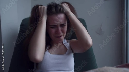 Young depressed woman pulling her hair with hands and crying sitting in chair by the window. Raining outside. Lonely woman in despair with suicide thoughts.