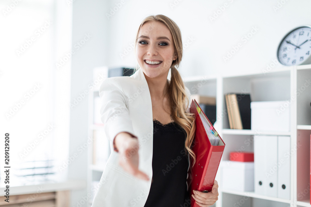 A young girl stands in the office next to the shelf, holds a folder with documents in her hand and stretches her right hand forward.