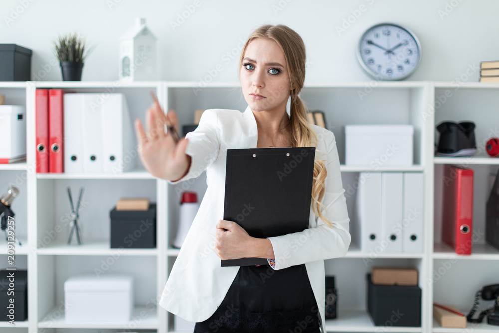 A young girl stands in the office next to the rack, holds documents and a pen in her hand and shows a stop sign. A young girl stands in the office next to the rack, holds documents and a pen in her hand and shows a stop sign.