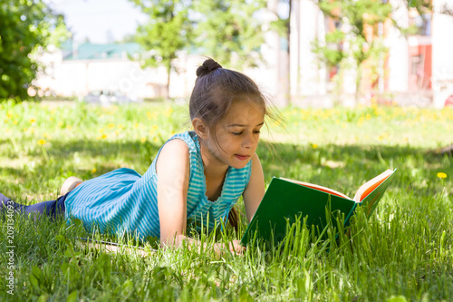 Cute pensive little girl lying on a green grass in summer city park and reading a book