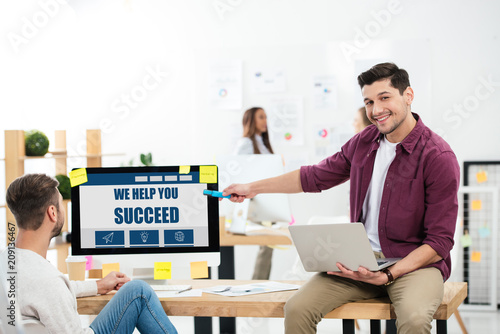 businessman pointing at we help you succeed inscription on computer screen while working at workplace with colleague in office