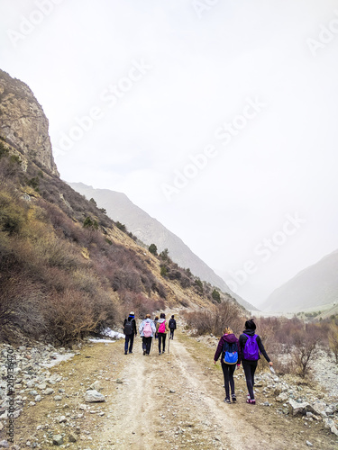 Group of hikers walking in mountains. A group of people with backpacks walking along the road. There are mountains from left. The sky is cloudy.