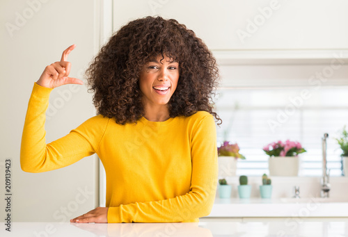 African american woman wearing yellow sweater at kitchen smiling and confident gesturing with hand doing size sign with fingers while looking and the camera. Measure concept.