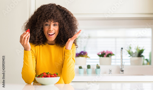 African american woman eating strawberries at home very happy and excited, winner expression celebrating victory screaming with big smile and raised hands