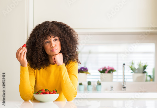 African american woman eating strawberries at home serious face thinking about question, very confused idea