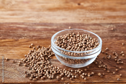 Glass bowl full of coriander seeds on rustic wooden background, close-up, side view, selective focus.
