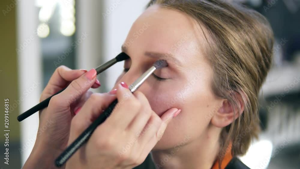 The professional make-up artist does make-up for the young cute model. Artist putting an eyes shadows using two brushes and does it together. Close up The professional make-up artist does make-up for the young cute model. Artist putting an eyes shadows using two brushes and does it together. Close up