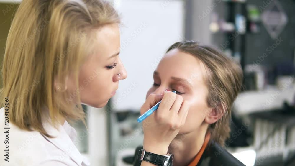 Make-up work. Make-up artist and model. Girl makeup with short fair hair. Makeup artist paints eyeliner lower eyelids model. Eyes makeup, studio Make-up work. Make-up artist and model. Girl makeup with short fair hair. Makeup artist paints eyeliner lower eyelids model. Eyes makeup, studio
