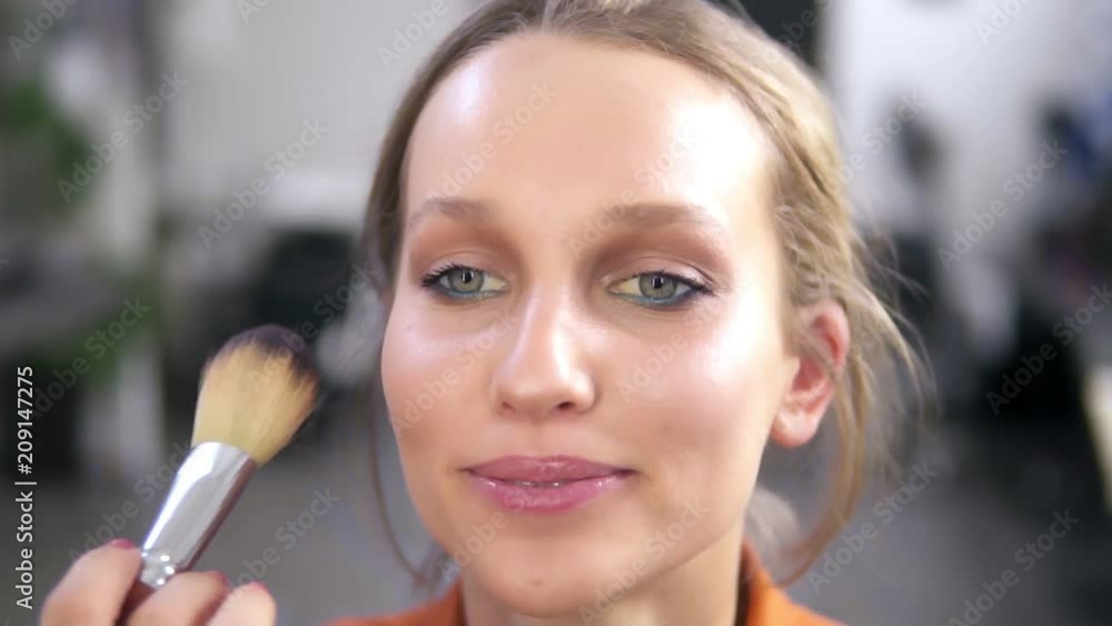 Front view of a beautiful smiling female model during the make up process. Make up artist completes the work, putting some powder using a brush. Close up footage Front view of a beautiful smiling female model during the make up process. Make up artist completes the work, putting some powder using a brush. Close up footage