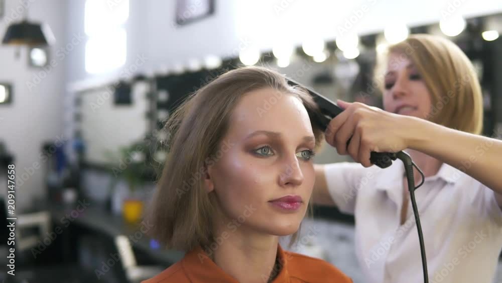 Attractive caucasian girl with make up in beauty salon. Hairdresser straightens her hair with an iron. Blurred background - white lamps and mirrors Attractive caucasian girl with make up in beauty salon. Hairdresser straightens her hair with an iron. Blurred background - white lamps and mirrors