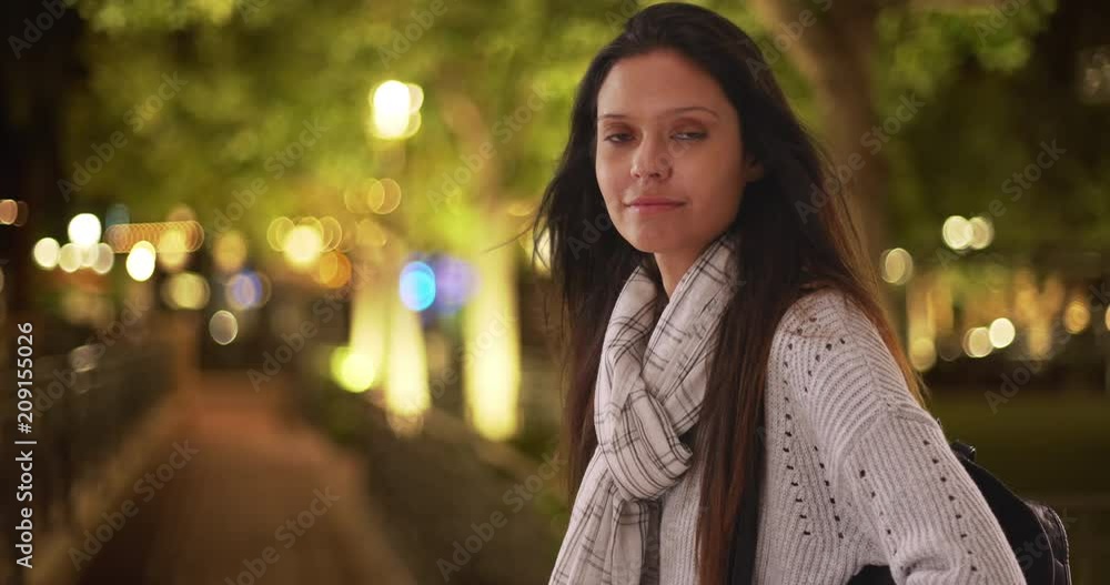 Beautiful female in striped scarf making eye contact while on city street at night, Caucasian woman with her backpack smiling and hair blowing in wind, 4k Beautiful female in striped scarf making eye contact while on city street at night, Caucasian woman with her backpack smiling and hair blowing in wind, 4k