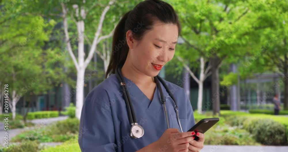 Close up of female millennial nurse dressed in scrubs and using smartphone to send texts on break outside hospital, Happy young Asian doctor texting with mobile device outdoors city clinic, 4k Close up of female millennial nurse dressed in scrubs and using smartphone to send texts on break outside hospital, Happy young Asian doctor texting with mobile device outdoors city clinic, 4k