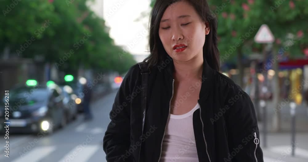 Chinese woman in black bomber jacket on city street with hair blowing in wind, Asian millennial tourist waits on sidewalk with pockets in Paris, 4k Chinese woman in black bomber jacket on city street with hair blowing in wind, Asian millennial tourist waits on sidewalk with pockets in Paris, 4k
