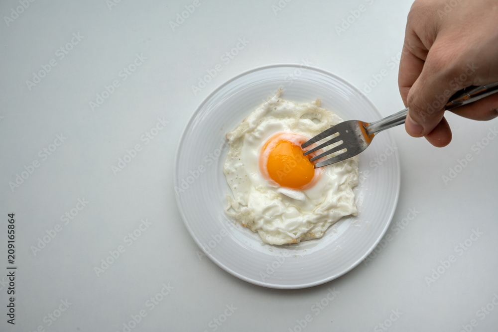 Hand holding fork to prick the egg yolk of beautiful fried egg in white plate isolated on white Hand holding fork to prick the egg yolk of beautiful fried egg in white plate isolated on white