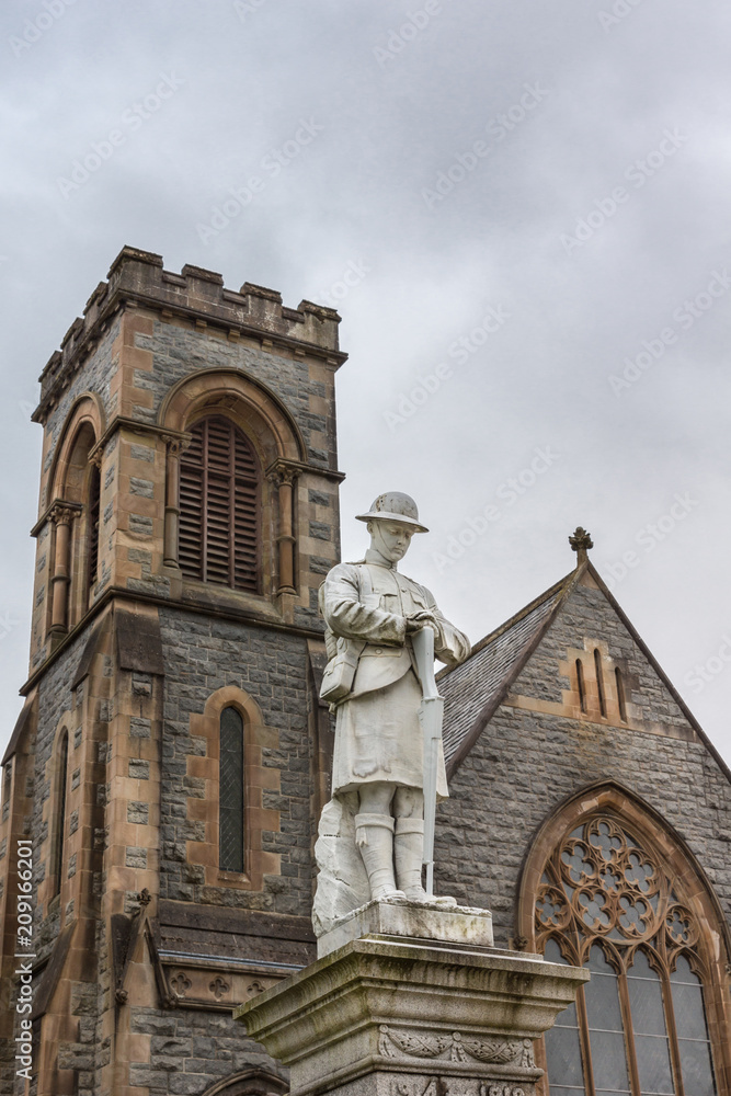 Fort William, Scotland - June 11, 2012: Closeup of white stone war memorial statue on The Parade against gray sky. Shows young soldier in kilt contemplating, Duncansburgh Church of Scotland in back.