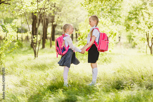 two little Schoolgirls in school uniform stand with backpacks and hold hands outdoors