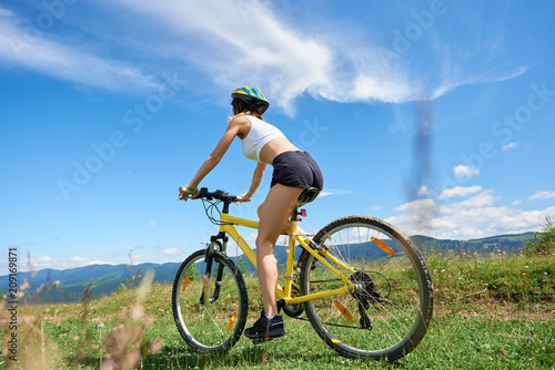 Wide angle view of active woman rider cycling on yellow mountain bike on a rural trail, against blue sky with clouds. Outdoor sport activity, lifestyle concept. Copy space