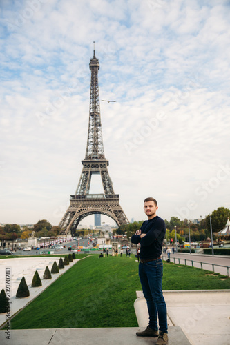 Handsome man wearing a dark blue pullover is standing on the background of the Eiffel Tower © Aleksandr