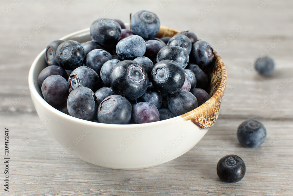Small bowl full of blueberries, some of them spilled on gray wood desk. Small bowl full of blueberries, some of them spilled on gray wood desk.
