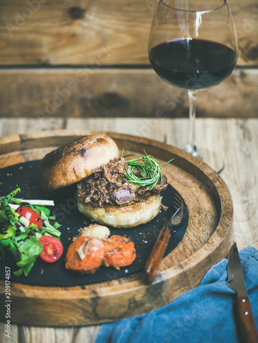 Pulled pork meat burger with vegetables and glass of red wine, wooden wall background, copy space