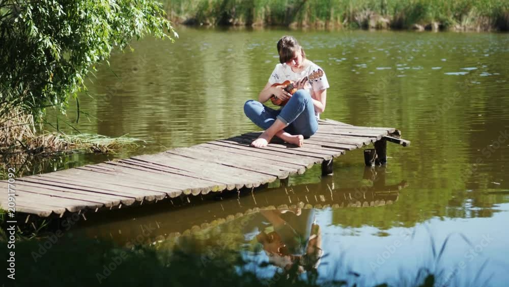 Young beautiful girl playing ukulele guitar sitting on the wooden pier on river at summer sunny day. Ukulele concept Young beautiful girl playing ukulele guitar sitting on the wooden pier on river at summer sunny day. Ukulele concept