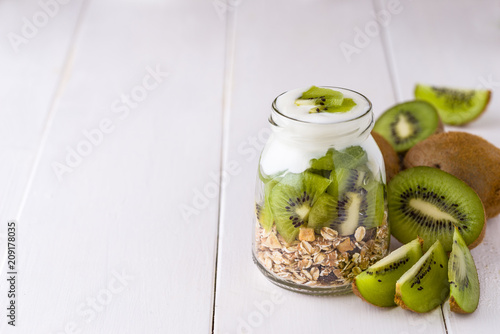 White yogurt with muesli in glass bowl with pieces of kiwi on top and whole pieces on the right side.