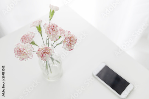 Top view of pink and white carnation flowers vase with smart phone on table in white room. Flat lay.