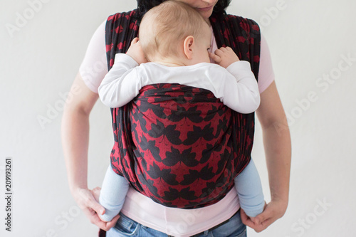 A cropped view of a baby in a woven wrap. A front view of a babywearing mother carrying her small child in a sling.