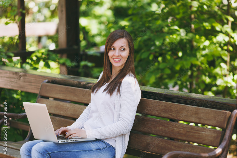 Young successful businesswoman in light casual clothes. Woman sitting on bench working on modern laptop pc computer in city park in street outdoors on nature. Mobile Office. Freelance business concept Young successful businesswoman in light casual clothes. Woman sitting on bench working on modern laptop pc computer in city park in street outdoors on nature. Mobile Office. Freelance business concept