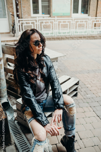 Casual beauty. Close up fashion woman, vintage denim biker leather jacket, rock  n roll style, fit girl posing on the street, stylish outfit. On the  background of light yard. Stock Photo