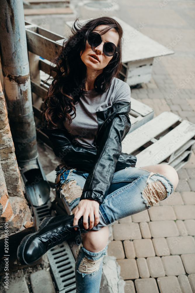Slice of summer goodness. Beautiful young woman in denim biker leather  jacket, rock n roll style, fit girl posing on the street, stylish outfit.  On the background of light yard. Stock Photo