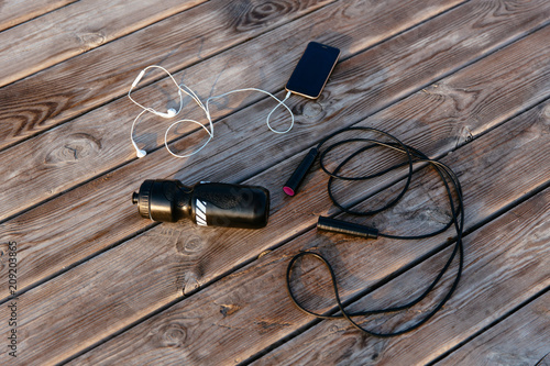 Photography Top view of smartphone with earphones, jump rope and bottle of water against the wood background