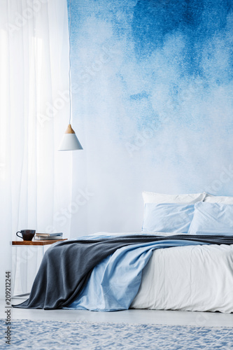 Bed with grey blanket against blue ombre wall in bedroom interior with lamp above table © Photographee.eu