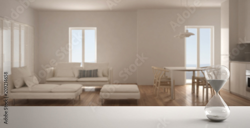 White table or shelf with crystal hourglass measuring the passing time over blurred empty space, living room with kitchen, architecture interior design, copy space background © ArchiVIZ