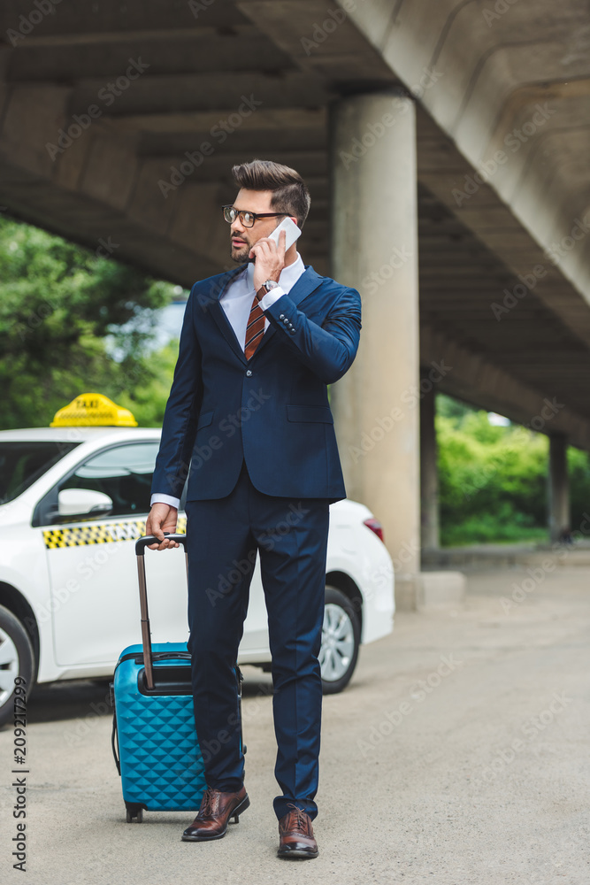 stylish young man talking by smartphone and looking away while standing with suitcase near taxi stylish young man talking by smartphone and looking away while standing with suitcase near taxi