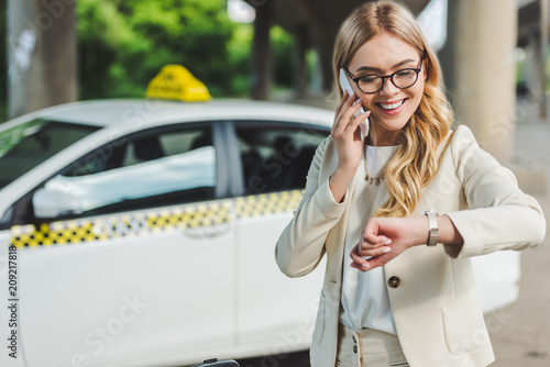 smiling blonde woman in eyeglasses talking by smartphone and checking wristwatch while standing near taxi cab © LIGHTFIELD STUDIOS