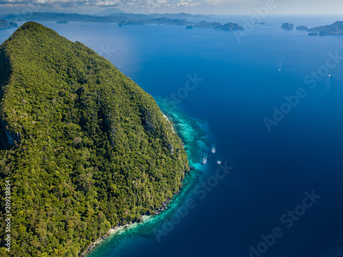 Aerial drone view of boats in a clear ocean, over a tropical coral reef, surrounded by towering jungle covered mountains