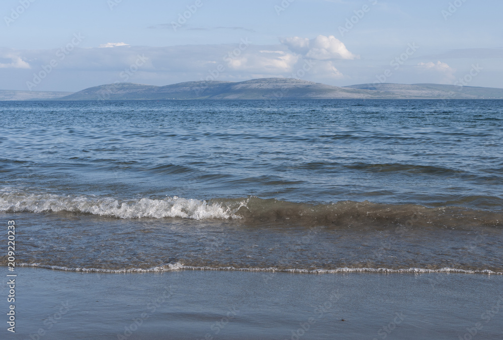 Waves crashing against a sandy beach on a sunny day, with tall hills in the background. Taken in Galway Bay in summer.