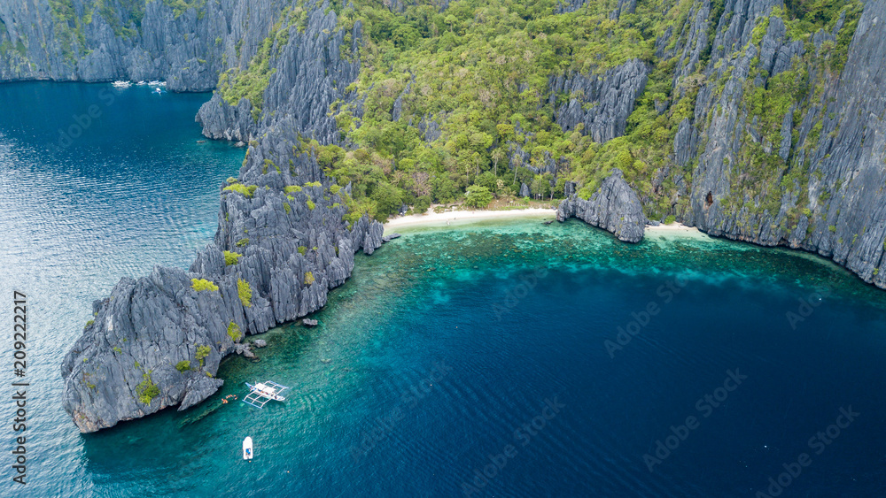 Aerial drone view of boats above a tropical coral reef and small sandy beach surrounded by huge cliffs (Secret Lagoon, Miniloc)