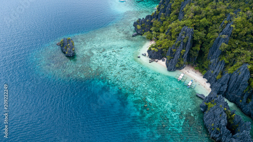 Aerial drone view of boats above a tropical coral reef and small sandy beach surrounded by huge cliffs (Secret Lagoon, Miniloc)