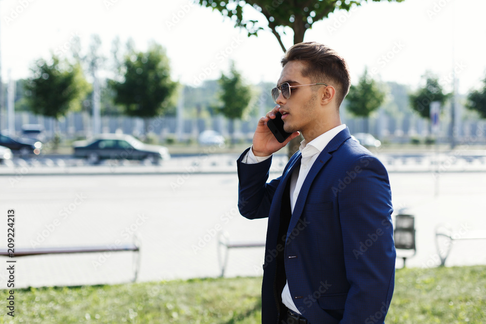 Handsome man in a business suit walks along the street in a sunny day and talks on his smartphone Handsome man in a business suit walks along the street in a sunny day and talks on his smartphone