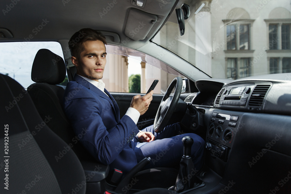 Smiling businessman sits insde the car and works with his smartphone