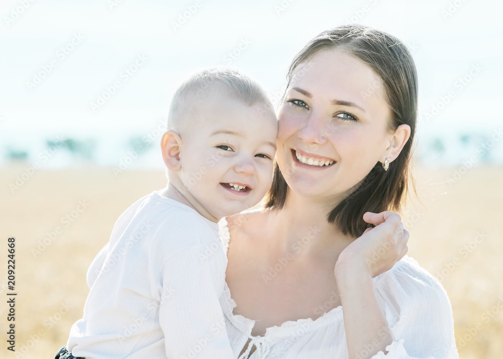 Fototapeta premium Family young happy pretty mother holding little baby son and smiling in golden wheat or rye field at sunny summer day. Blurred skyline background. Concept simple rustic life childhood and motherhood