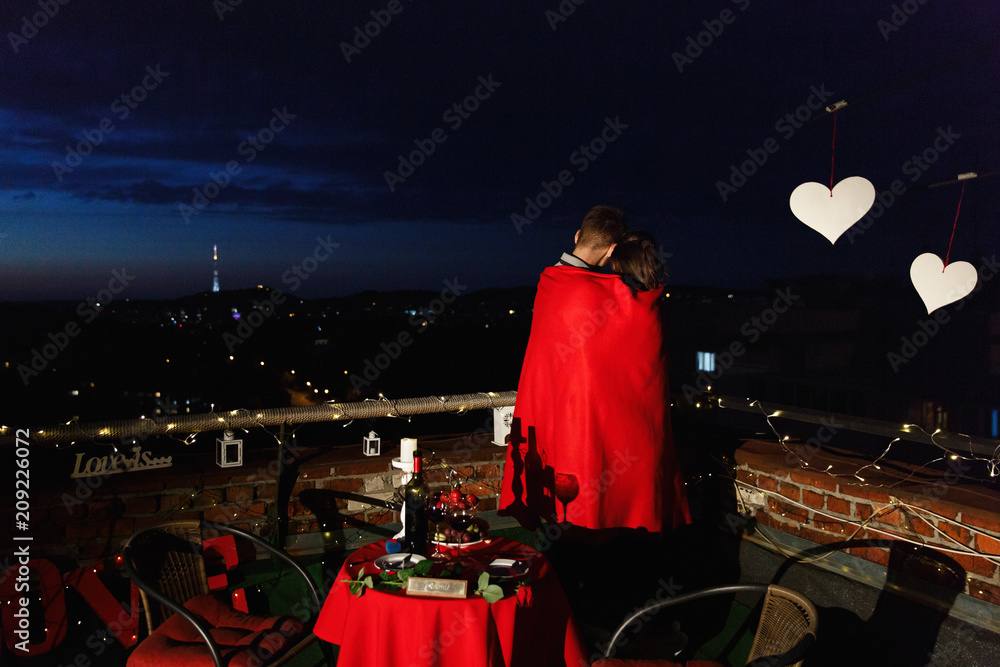 Boy and girl hug each other tender standing on the rooftop in the rays of evening lights and having a romantic dinner Boy and girl hug each other tender standing on the rooftop in the rays of evening lights and having a romantic dinner