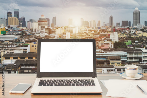 Mockup image of laptop with blank white screen,smartphone,coffee cup on wooden table view outdoors of office building dense cityscape background.