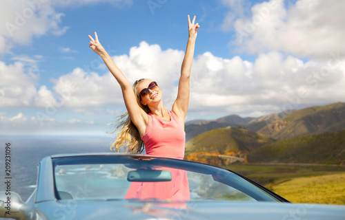 travel, summer holidays, road trip and people concept - happy young woman wearing sunglasses in convertible car showing peace sign over bixby creek bridge on big sur coast of california background