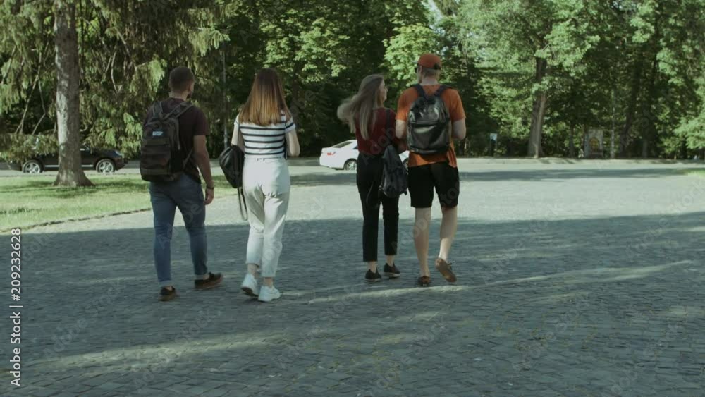Rear view of college friends with backpacks walking and communicating right after lecture. Happy university students walking in campus after study. Steadicam stabilized shot. Rear view of college friends with backpacks walking and communicating right after lecture. Happy university students walking in campus after study. Steadicam stabilized shot.