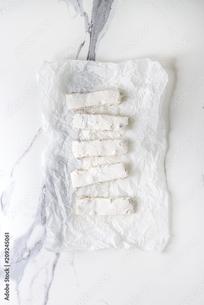 Homemade honey nuts nougat turron sliced on crumpled paper with sugar powder over white marble background. Flat lay, space. Homemade honey nuts nougat turron sliced on crumpled paper with sugar powder over white marble background. Flat lay, space.