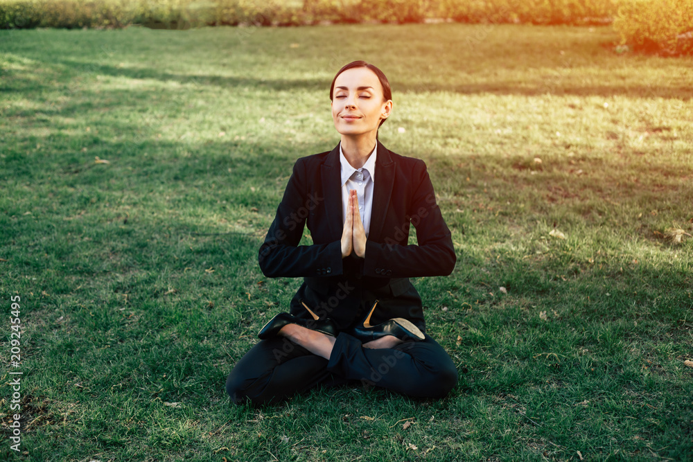 Young beautiful modern business woman in a suit meditating in a park sitting in a lotus position in the fresh air Young beautiful modern business woman in a suit meditating in a park sitting in a lotus position in the fresh air